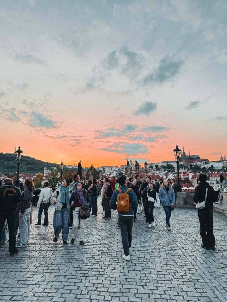 Crowd of tourists walking on the cobblestone Charles Bridge in Prague at sunset, with a stunning orange and pink sky over the city and Prague Castle in the distance.