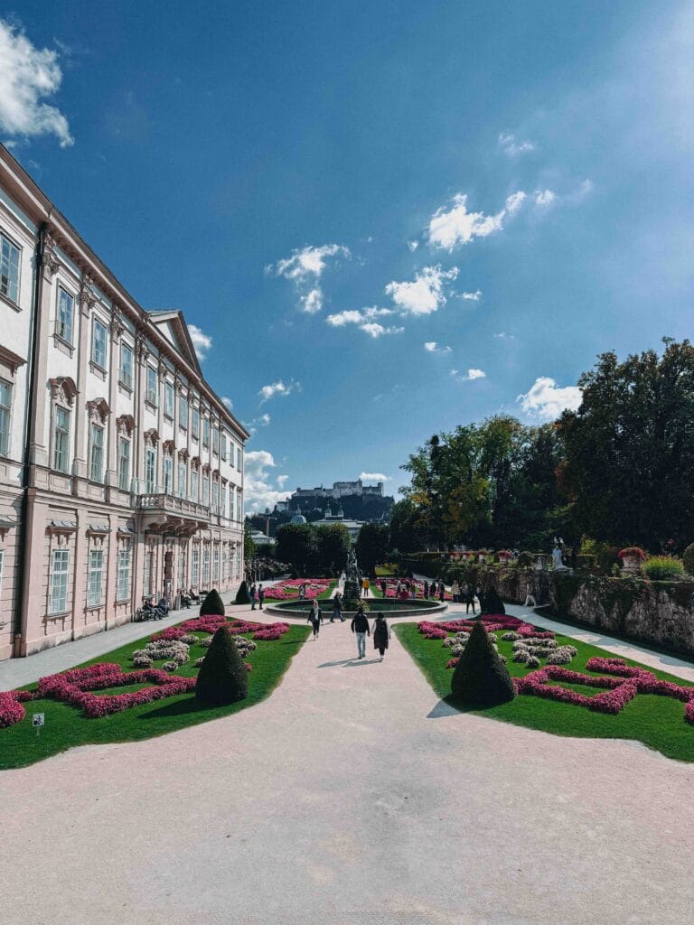A wide view of the meticulously maintained Mirabell Gardens in Salzburg, showing pink and white flowers lining the path, the baroque palace to the left, and Hohensalzburg Fortress visible in the distance.