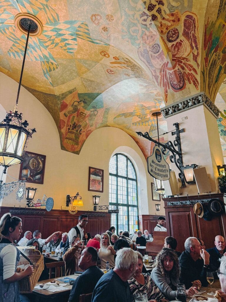 Interior of Hofbräuhaus München dining hall with painted ceilings, wooden tables, and "Stammtisch" signs.