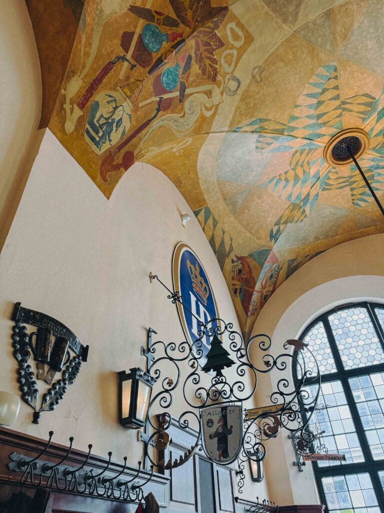 Close-up of ornate painted murals and iron chandeliers on the vaulted ceilings of Hofbräuhaus.