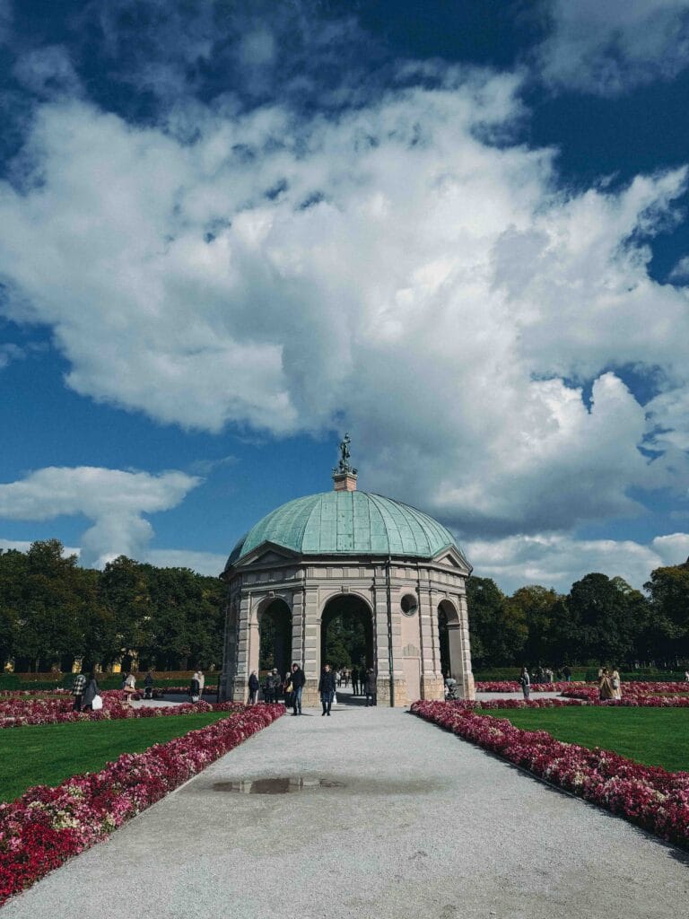 The octagonal, domed Temple of Diana pavilion in the center of the Hofgarten in Munich, surrounded by green lawns and beds of red flowers, under a dramatic cloudy sky.