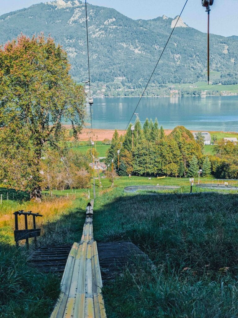 A summer toboggan run descending a grassy hill towards Lake Wolfgangsee, with mountains and green fields in the background, showing a person preparing to ride.