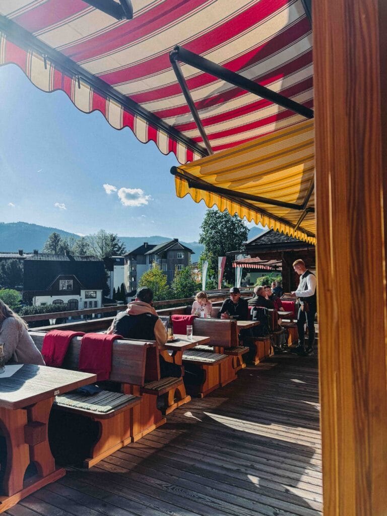 Guests dining on the sunny outdoor wooden terrace of Dorf-Alm zu St. Wolfgang, shaded by a red-and-white striped awning, with a waiter taking an order and mountain views in the distance.