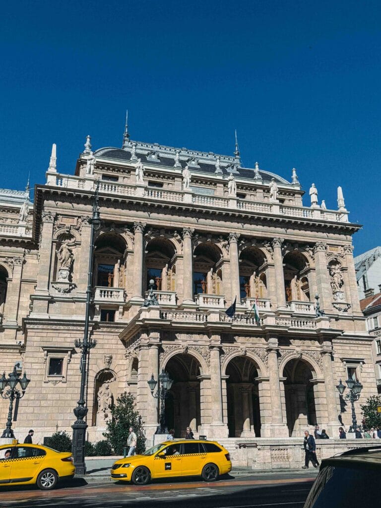 The neo-Renaissance exterior of the Hungarian State Opera House in Budapest on a clear day, with yellow taxi cabs parked in front.