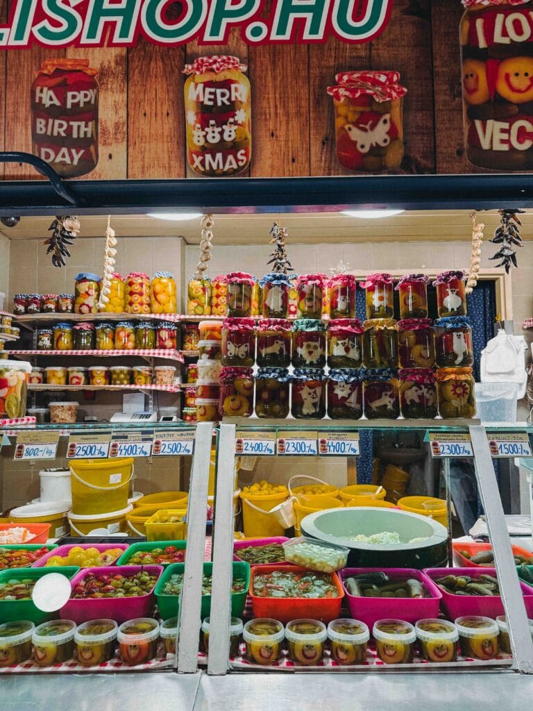 Stall filled with colorful jars of Hungarian pickled vegetables and preserves, including cucumbers, peppers, and fruit, inside the Great Market Hall in Budapest.