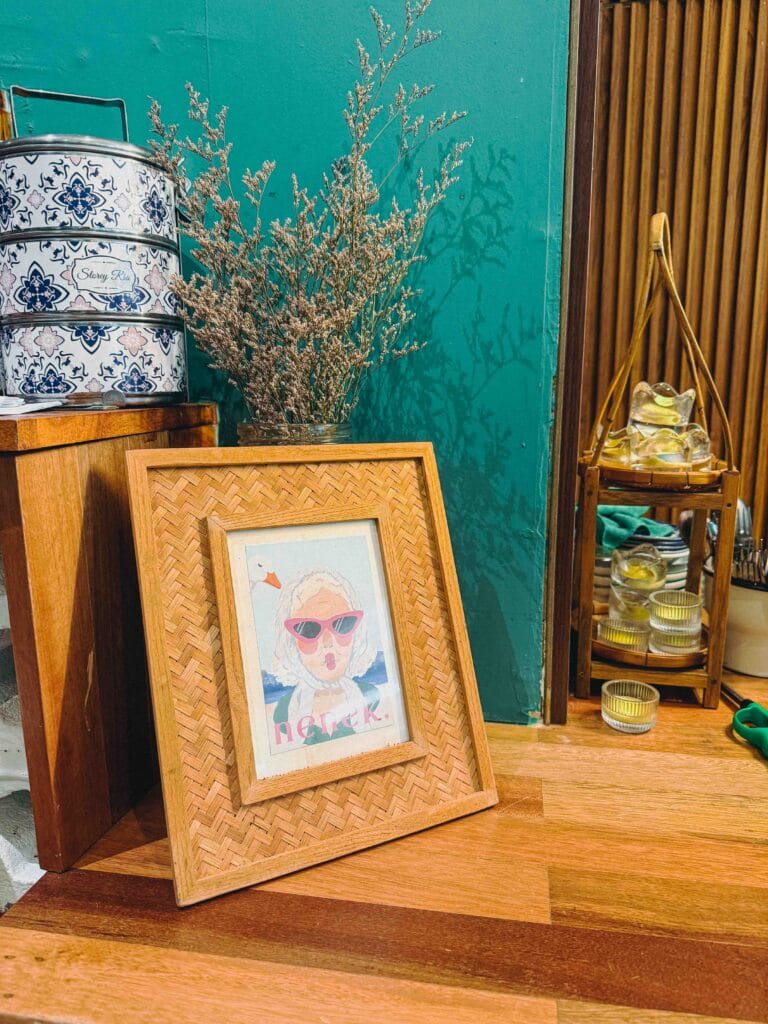 Rattan-framed nenek print next to a blue and white Peranakan-pattern tiffin carrier and dried flowers at Nenek Johor Bahru