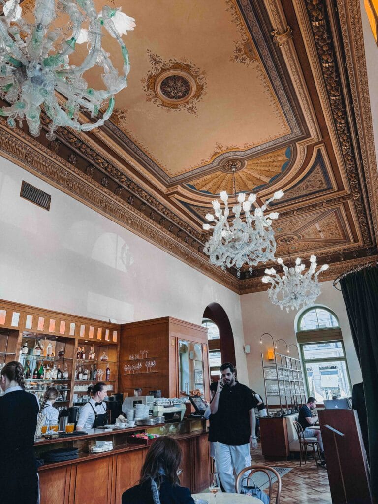 Interior view of Café Savoy Prague featuring the stunning Neo-Renaissance ceiling mural, towering white walls, and ornate crystal chandeliers over the bar area.