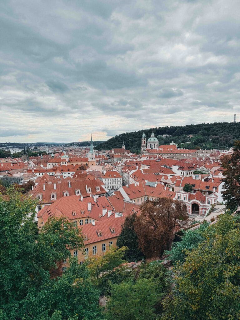 Panoramic view over the red tiled rooftops of Prague's Lesser Town (Malá Strana) with St. Nicholas Church and a forested hill in the distance.