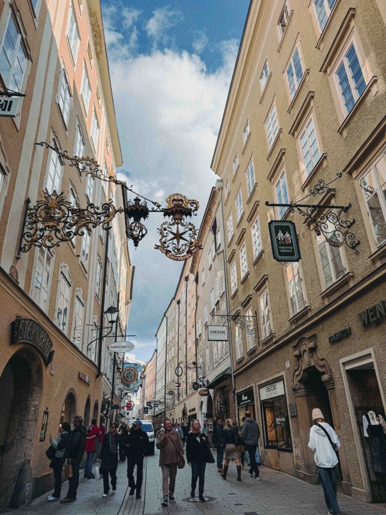 The famous Getreidegasse street in Salzburg, lined with tall historic buildings and featuring ornate, wrought-iron guild signs hanging above the heads of pedestrians.