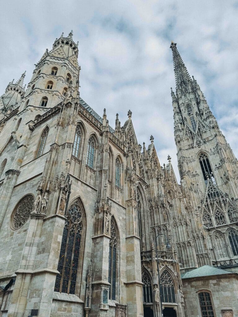 A dramatic, upward-looking view of the exterior of St. Stephen's Cathedral in Vienna, highlighting the intricate Gothic architecture and the tall, spired South Tower against the sky.