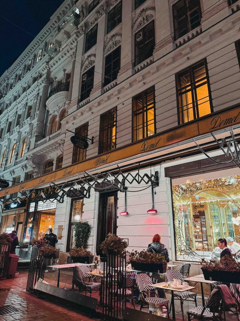 The elaborate exterior of Café Demel in Vienna at night, showing the outdoor seating area and the illuminated window displays of pastries.