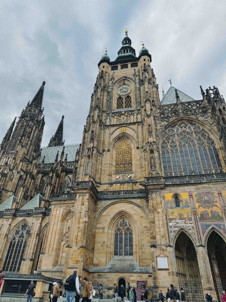 A low-angle, upward view of the ornate Gothic facade of St. Vitus Cathedral in Prague Castle, highlighting the main tower, the clock, and the golden mosaic of "The Last Judgement" above the portal.