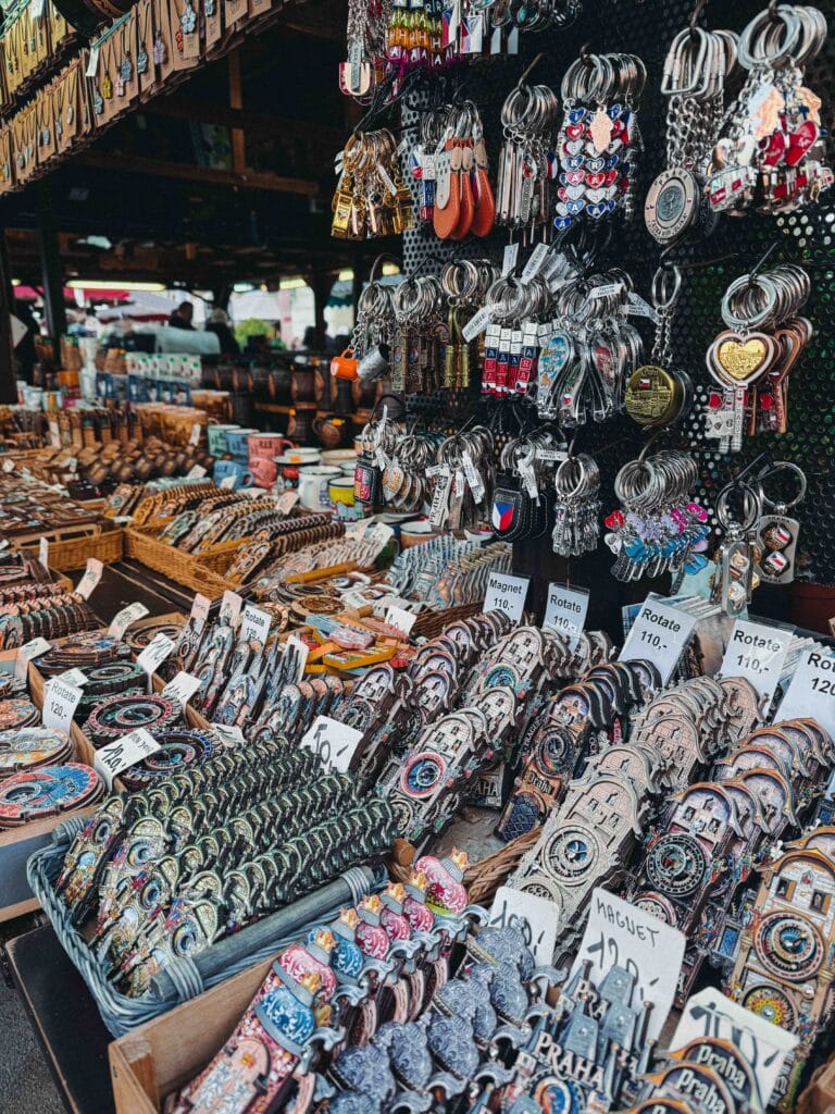 A souvenir stall at Havel's Market in Prague displaying rows of decorative magnets, keychains, and small wooden trinkets, with prices listed in Czech Koruna.