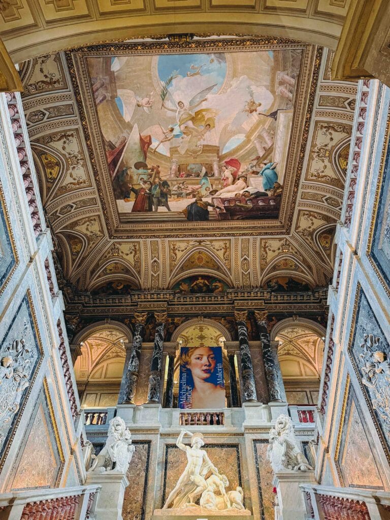 The opulent interior of the Kunsthistorisches Museum, showing the main entrance hall with a marble staircase, ceiling frescoes, and gilded decoration.