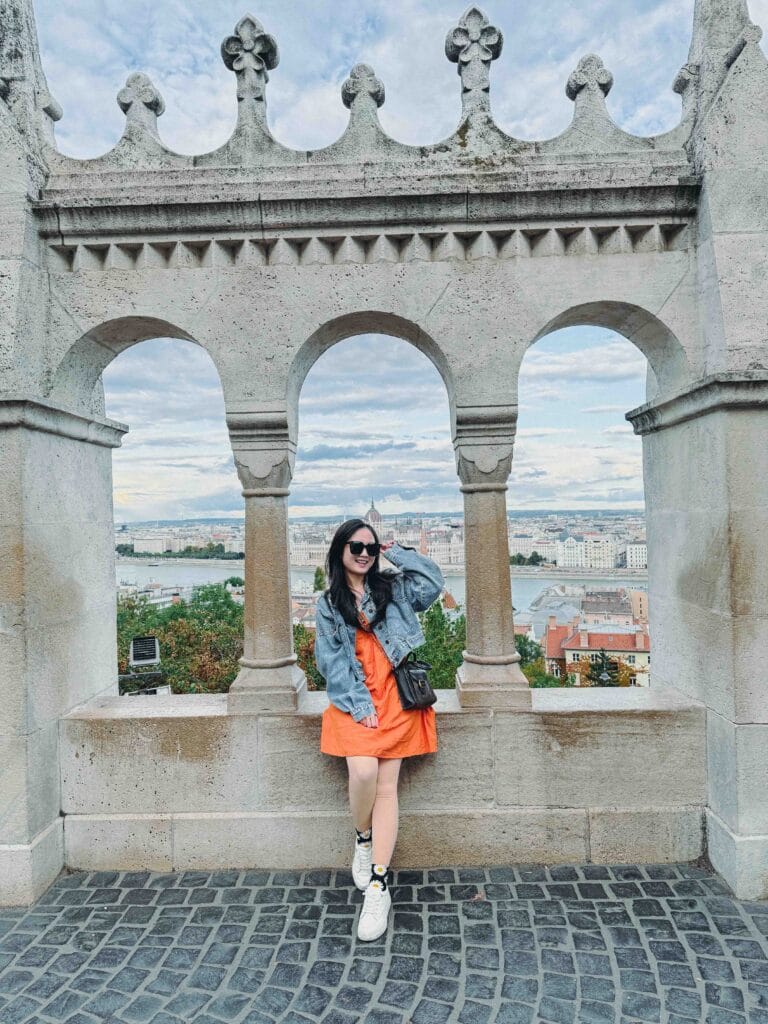 A panoramic view of the Pest side of Budapest from Fisherman's Bastion, showing the Danube River, the Hungarian Parliament Building, and the Chain Bridge, Valerie leaning on the railing, viewing the city.