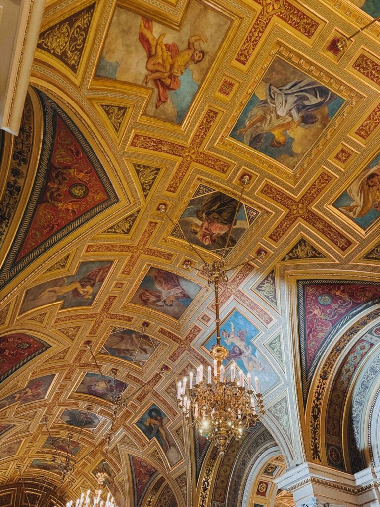 Highly ornate, gilded, and frescoed ceiling of the Hungarian State Opera House with classical paintings and a crystal chandelier hanging from the center.