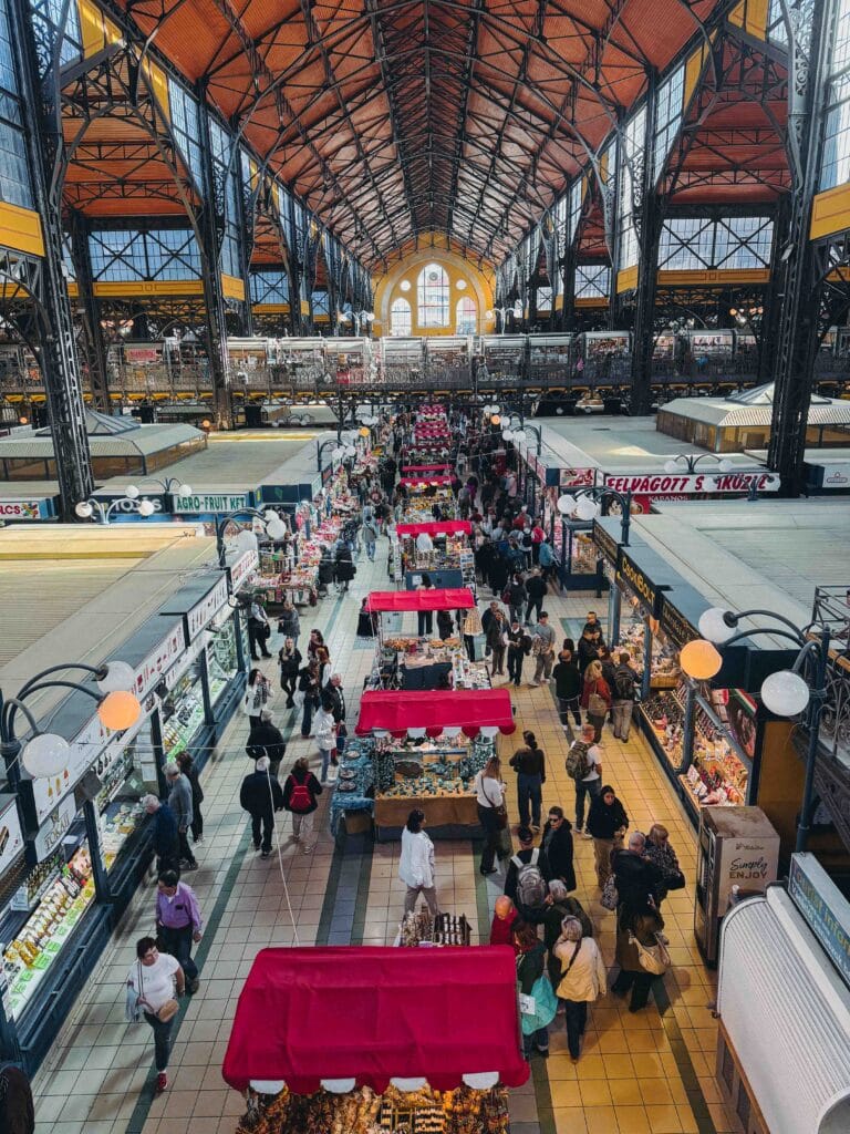 Panoramic interior view of the Great Market Hall in Budapest, showing the immense arched ceiling, upper floor food stalls, and strings of hanging Hungarian paprika.