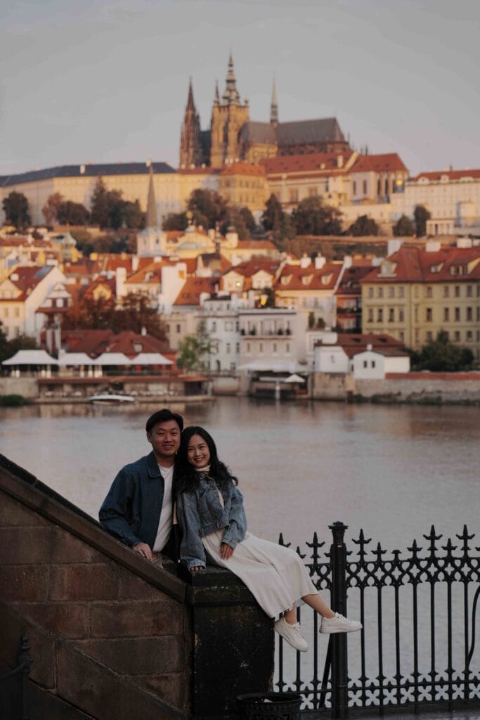 Li Yang and Valerie smiling and posing together on the Charles Bridge in Prague at sunset, with the Vltava River and the panoramic skyline of Prague Castle in the background.