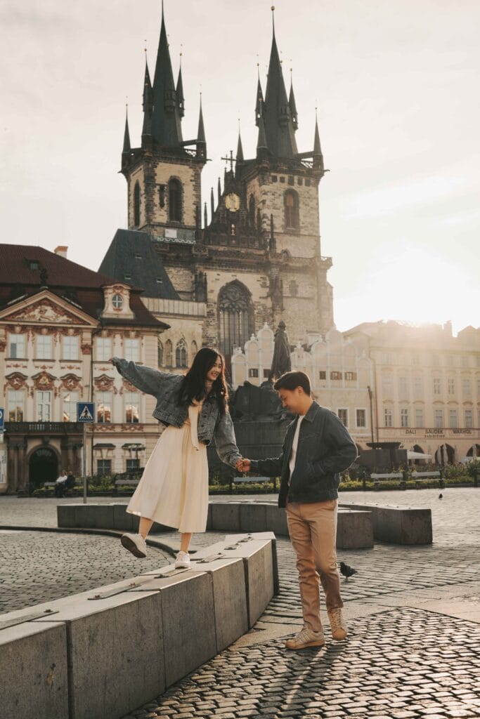 Li Yang and Valerie holds hands in Prague's Old Town Square, with the woman balancing on a ledge, and the twin Gothic towers of the Church of Our Lady before Týn rising dramatically in the background.