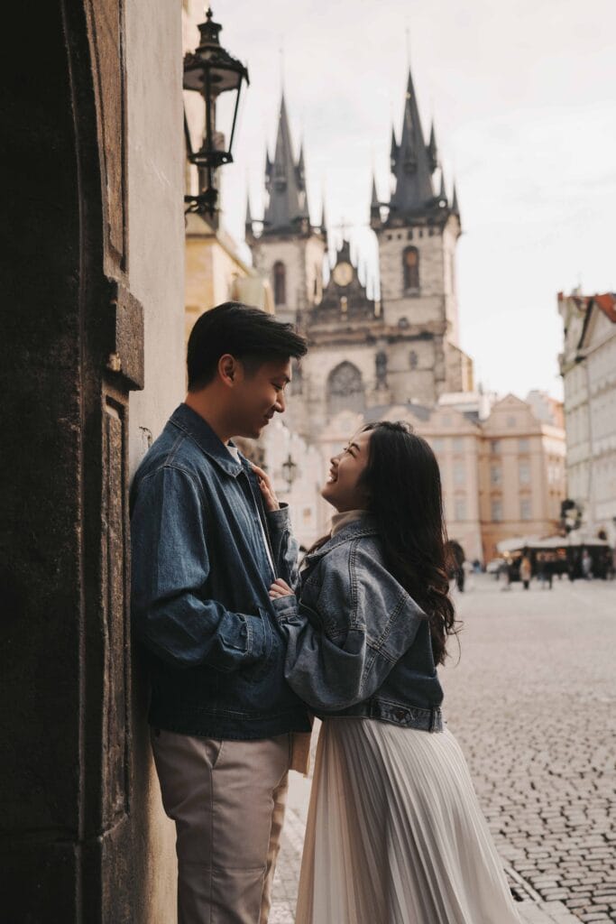 A romantic close-up of Li Yang and Valerie smiling at each other in a cobblestone alley in Prague, with the Gothic towers of the Church of Our Lady before Týn softly blurred in the background.