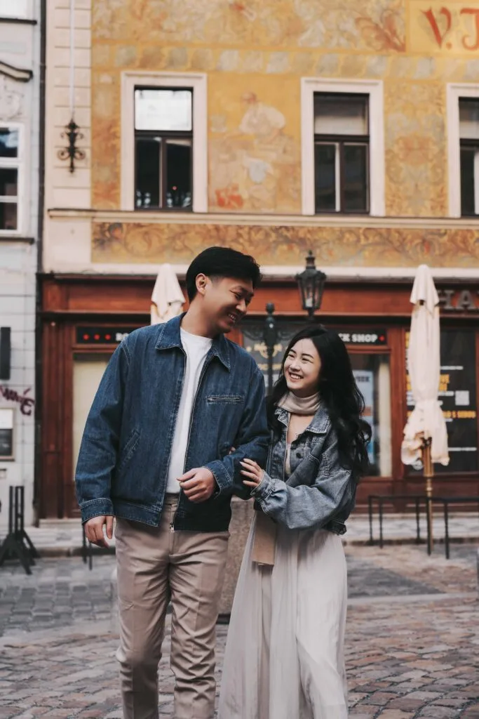Li Yang and Valerie walking arm-in-arm and laughing on the cobblestone streets of Prague Old Town with a historic frescoed building behind them
