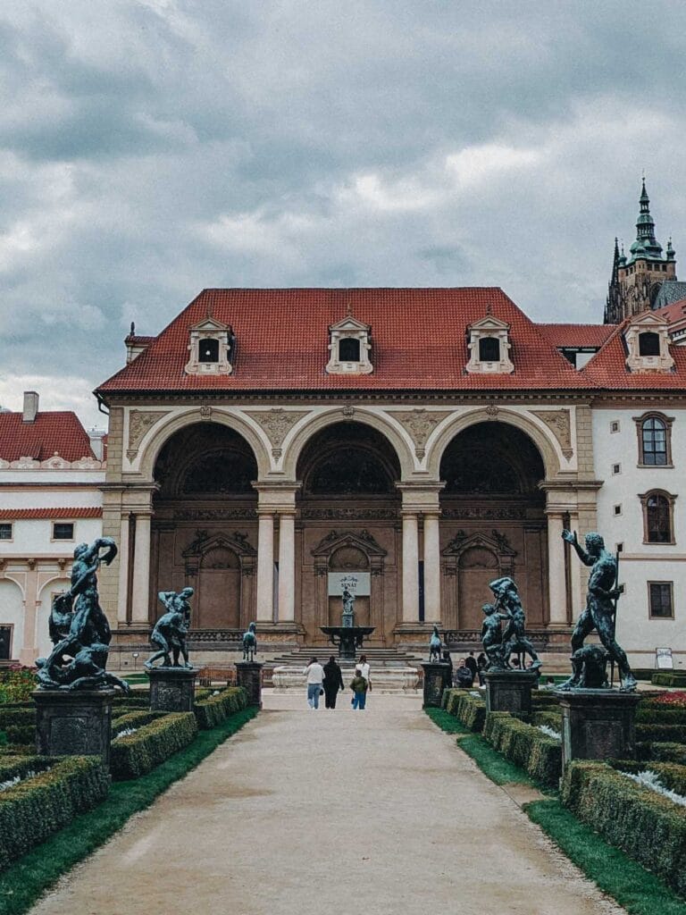 The impressive Sala Terrena façade of Wallenstein Palace in Prague, viewed from the gardens, with a path lined by bronze statues and hedges.