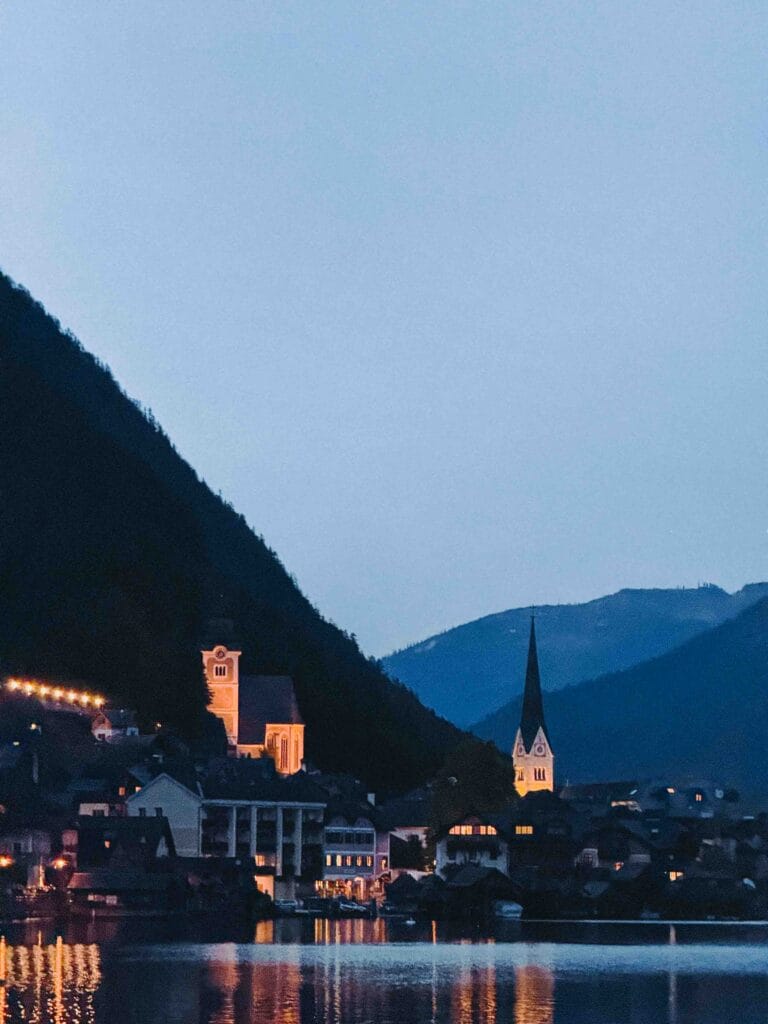 Iconic night view of Hallstatt, Austria, showing the illuminated houses and church spires reflecting in Lake Hallstatt, framed by dark mountains.