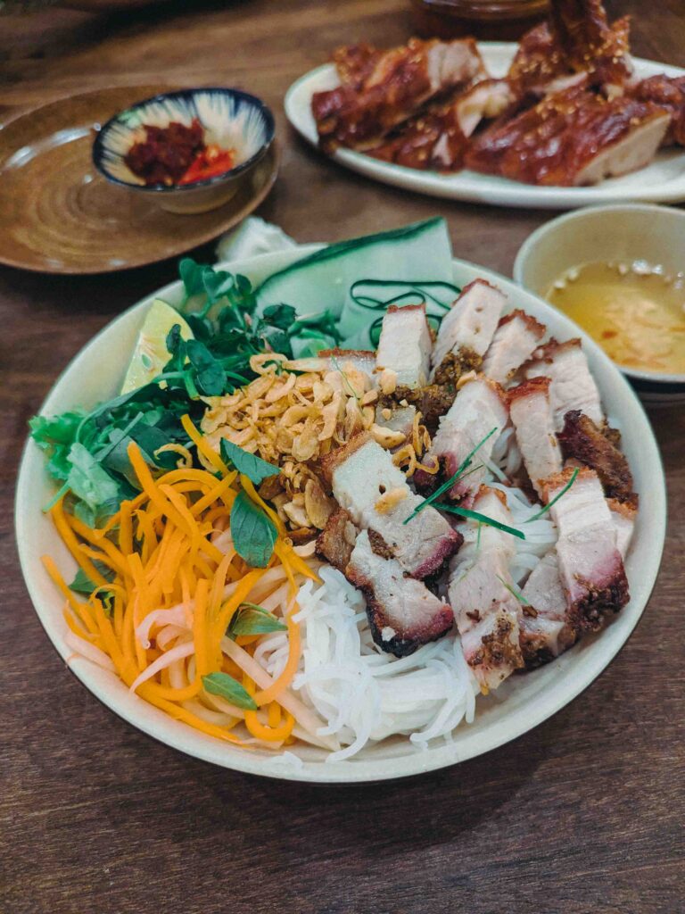 Roasted pork with fresh rice noodles, pickled carrot and daikon, fresh herbs and chilli dipping sauce in a bowl at Cơm Linh Hội An