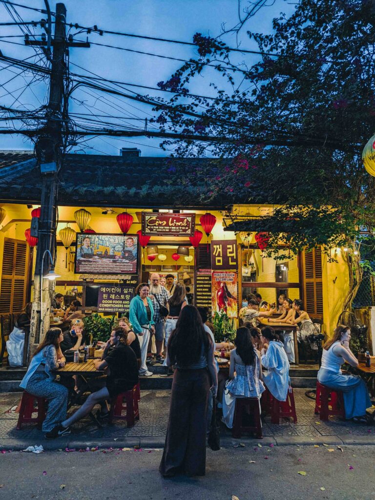 Cơm Linh restaurant exterior at night in Hoi An with red lanterns yellow walls and outdoor diners on low stools