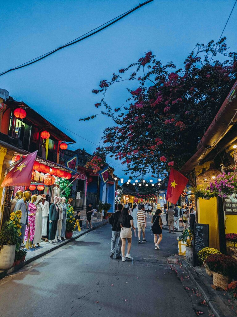 Hoi An Old Town street at dusk with red lanterns Vietnamese flags bougainvillea trees tailor shop mannequins and tourists walking