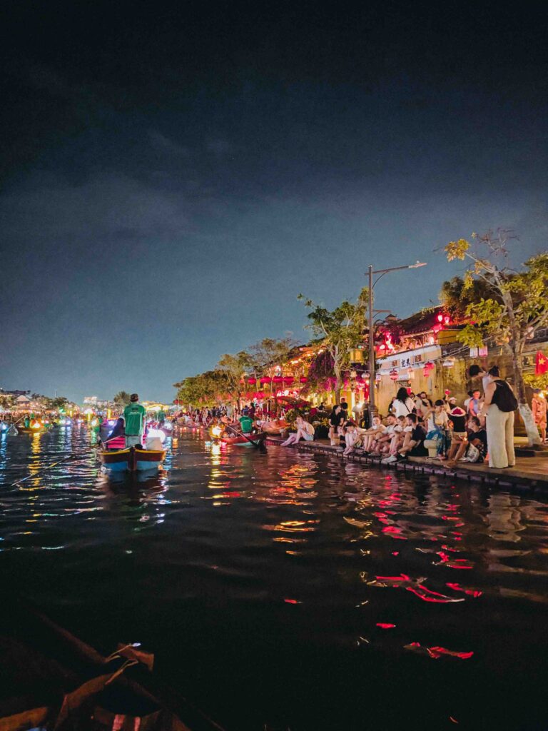 Lantern boats glowing on the Thu Bon River at night in Hoi An with lit shophouses and crowds sitting along the riverbank