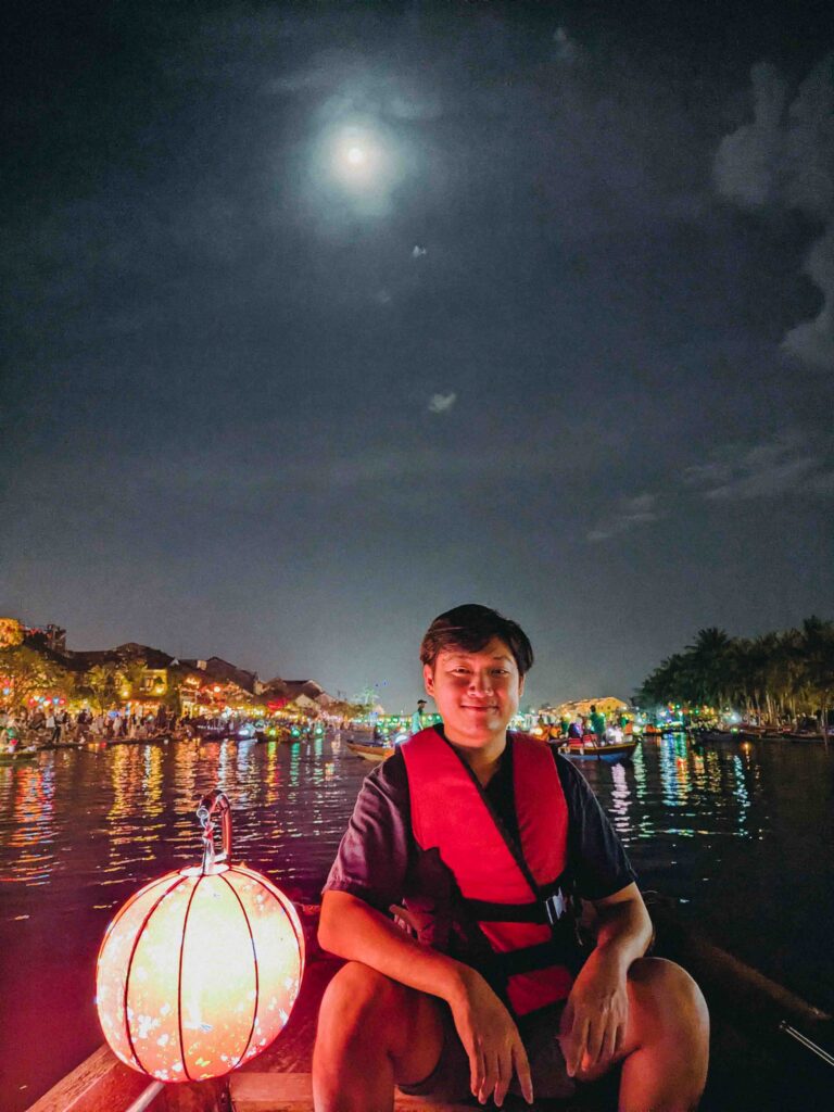 Li Yang in life jacket sitting in a lantern boat on the Thu Bon River in Hoi An at night with full moon above and colourful city lights reflected on the water