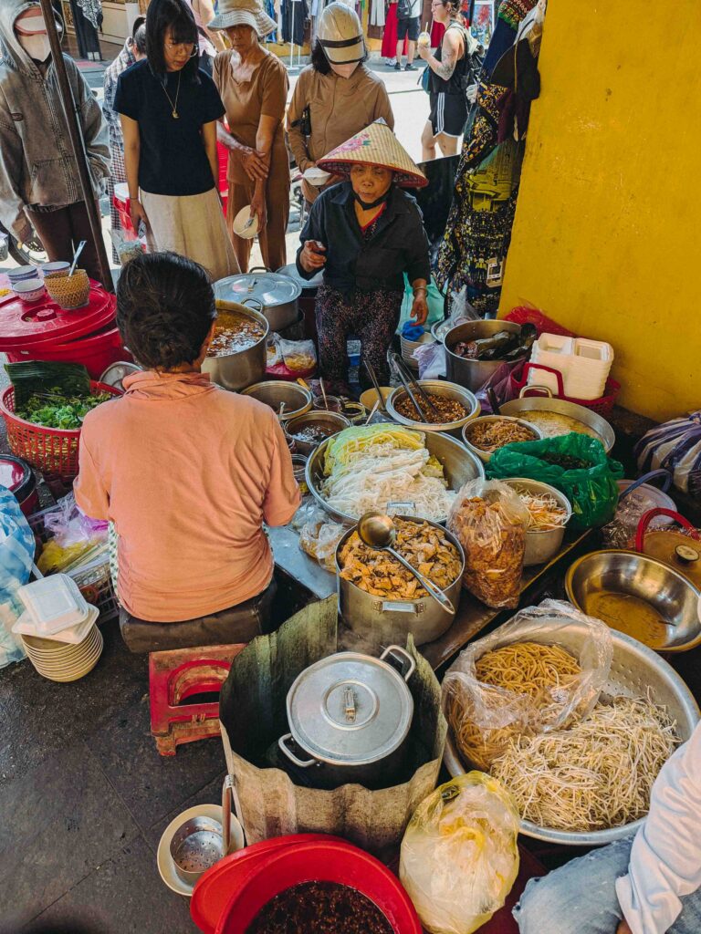 Vietnamese street food vendor in conical hat beside large pots of vegetarian dishes and tofu on a Hoi An Old Town street on the 15th lunar calendar day