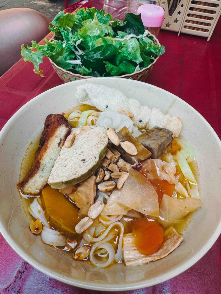 Bowl of Vietnamese vegetarian noodles with tofu fish cake peanuts and fresh herb basket eaten on a Hoi An Old Town street on the 15th lunar day