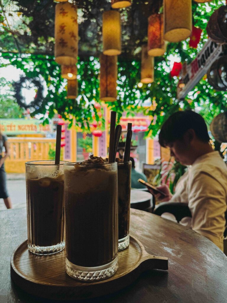 Three glasses of iced Vietnamese coffee on a wooden tray at Dudu Cafe Hoi An with green tree canopy and Chinese New Year decorations