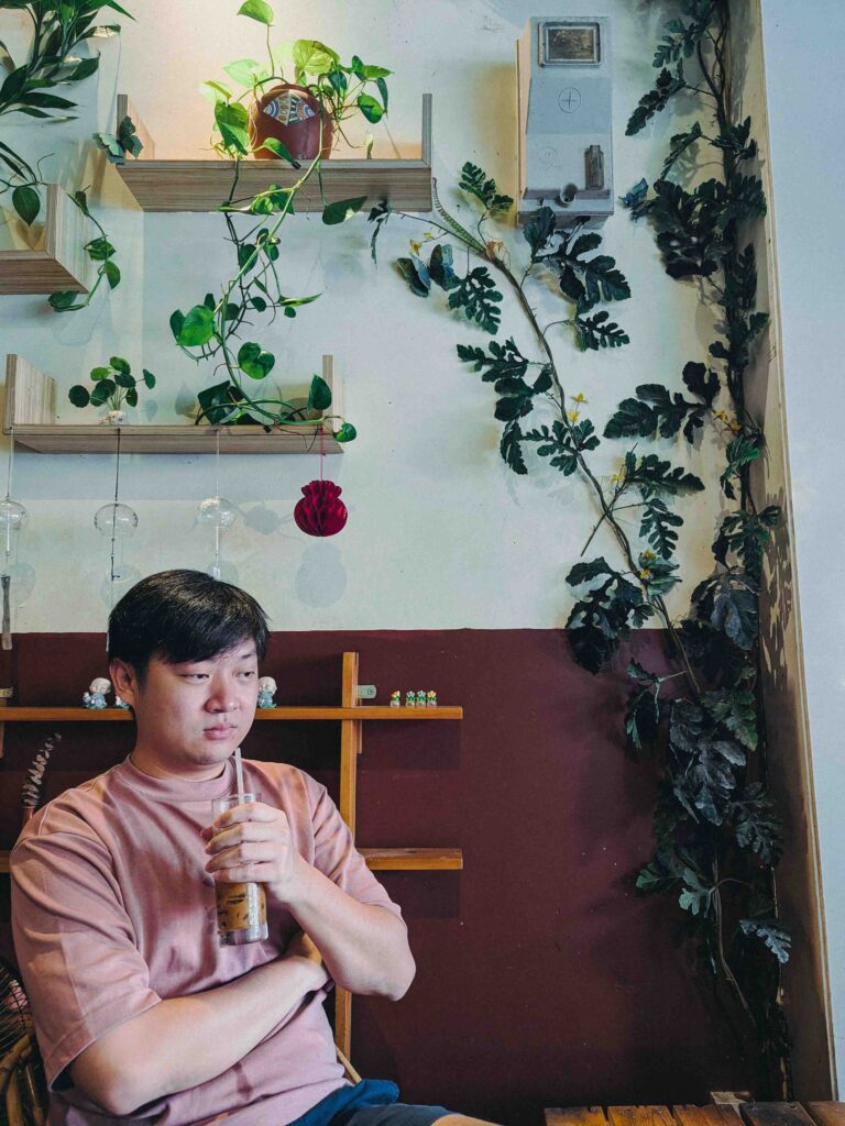 Li Yang holding iced coffee inside Mun Café Da Nang with green trailing plants on the wall