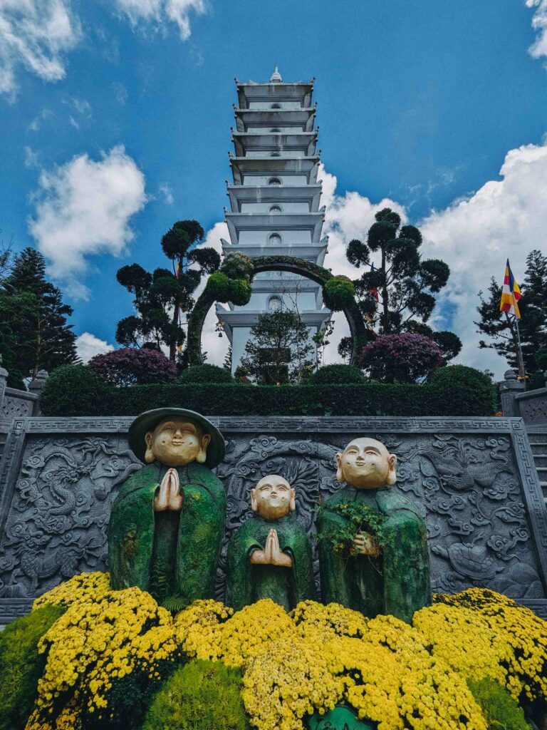 Three green monk statues with yellow chrysanthemums in front of white multi-tiered pagoda at Linh Phong Temple Ba Na Hills