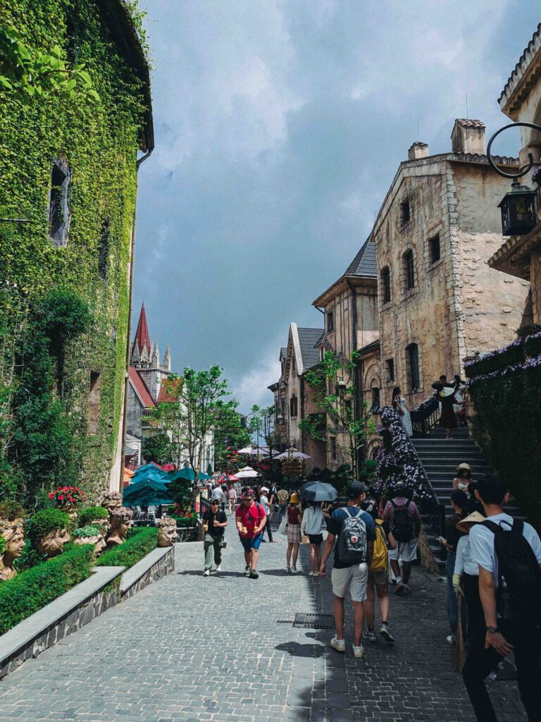 Tourists walking along cobblestone street of French Village at Ba Na Hills with ivy-covered walls and European-style buildings