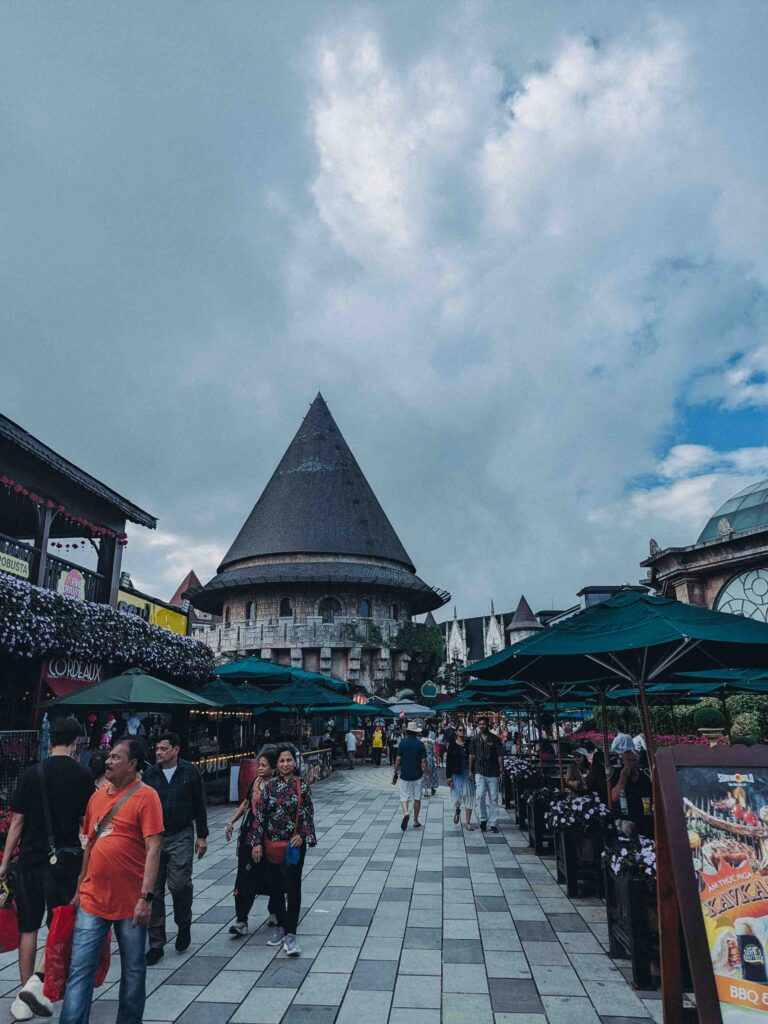 Tourists walking through the French Village town square at Ba Na Hills with a conical stone tower and outdoor dining umbrellas
