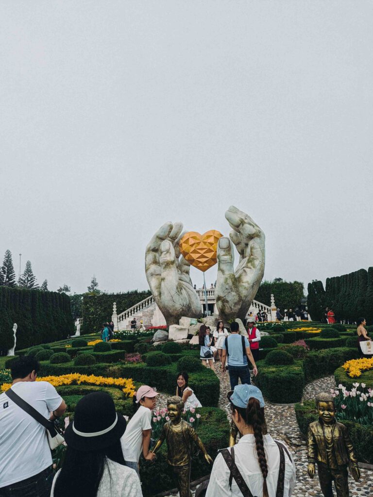 Giant stone hands holding a golden heart sculpture in the flower garden at Ba Na Hills with topiary hedges and tulips