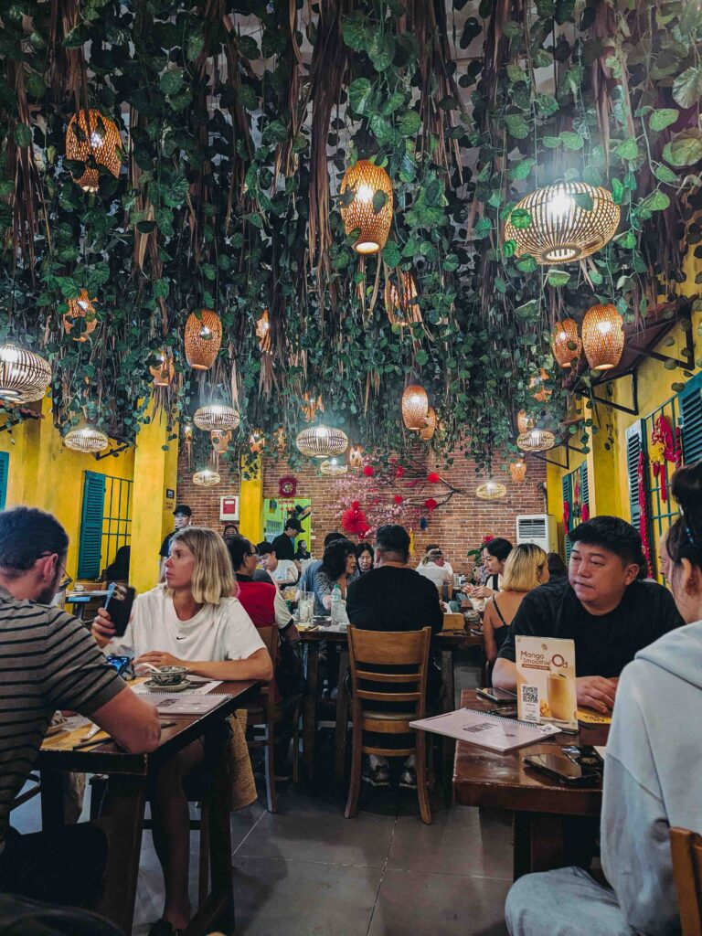 Lively dining room at Ăn Thôi Da Nang with yellow walls, hanging rattan lanterns, and trailing vines above packed tables