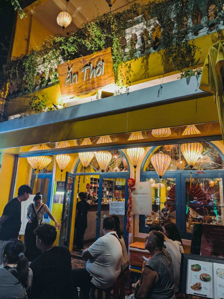 An Thôi Michelin-listed restaurant exterior at night in Da Nang with silk lanterns trailing vines and crowds waiting outside