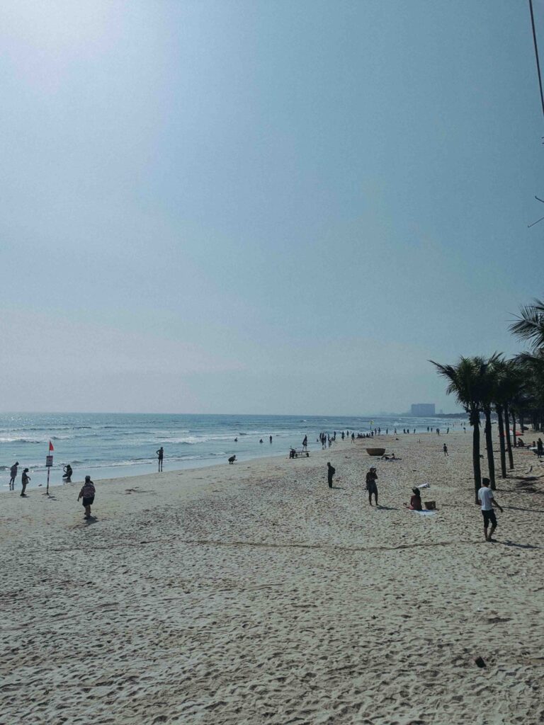 Da Nang Beach wide white sand shoreline with people walking along the water's edge palm trees and calm blue sea on a sunny day