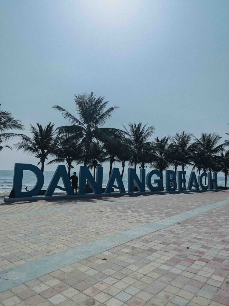 Large Da Nang Beach landmark sign with man standing beside it coconut palm trees and the South China Sea in the background