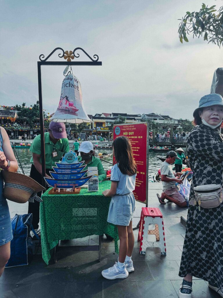 Official lantern boat ticket counter beside the Thu Bon River in Hoi An with staff in green uniforms and boat-shaped price display