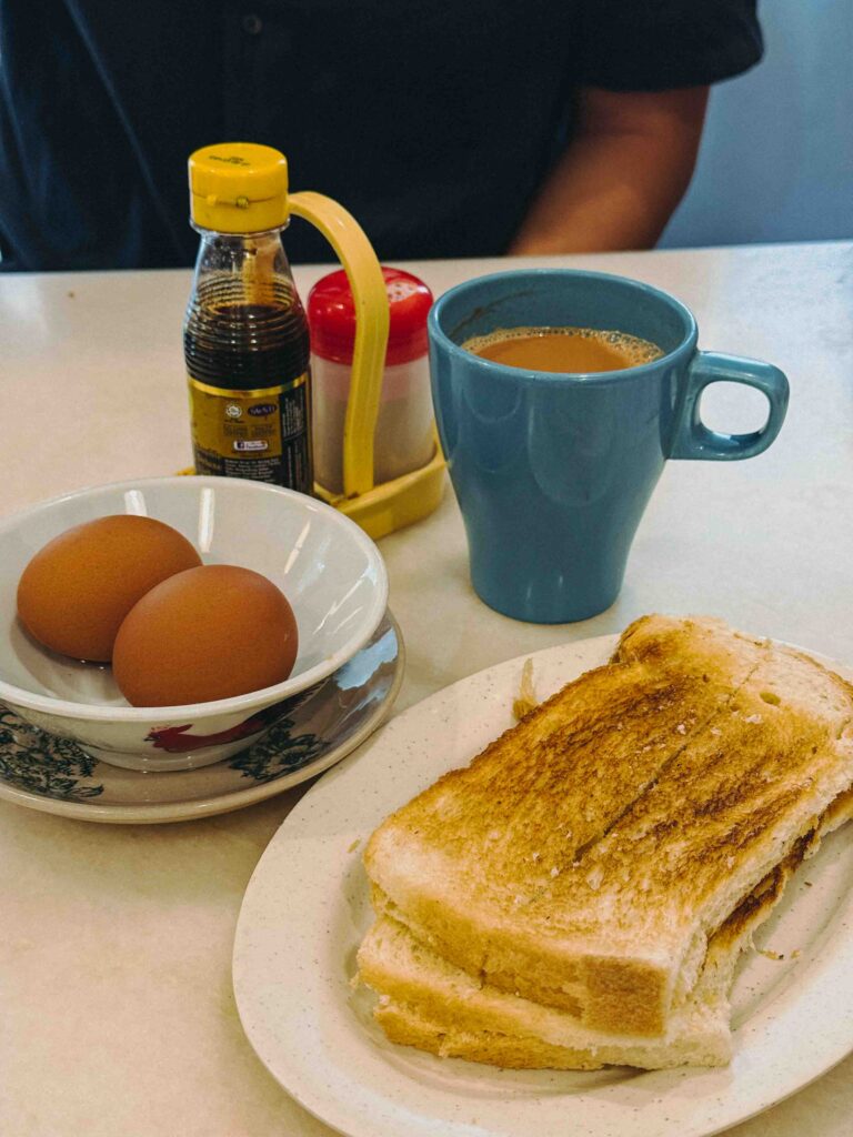 Village Park Coffee Set with roti bakar, half boiled eggs in bowl, and kopi in blue mug on white table